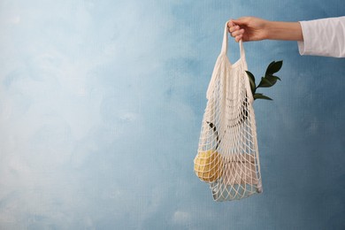 Woman holding net bag with different items against light blue wall, closeup and space for text. Conscious consumption