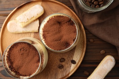 Photo of Delicious tiramisu in glasses, coffee beans and cookies on wooden table, flat lay