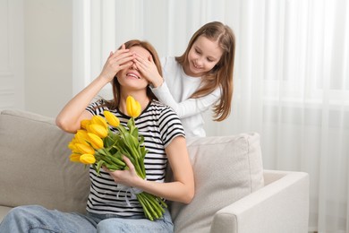Photo of Daughter covering mother's eyes with her palms and congratulating with bouquet of yellow tulips at home, space for text