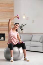 Young woman doing exercise with dumbbells on fitness ball at home