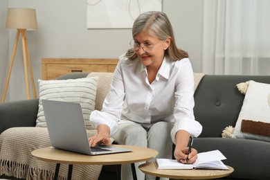 Beautiful senior woman writing something in notebook while using laptop at home