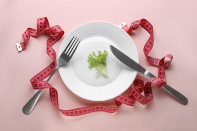 Plate with lettuce, measuring tape and cutlery on pale pink background, flat lay. Diet concept