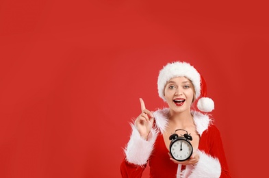 Beautiful Santa girl with alarm clock on red background, space for text. Christmas eve