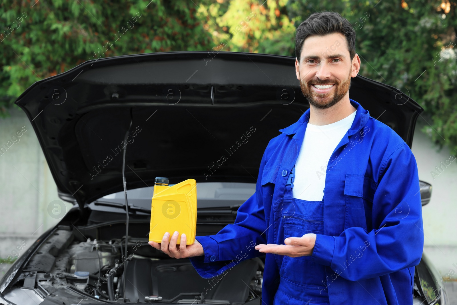 Photo of Smiling worker showing yellow container of motor oil near car outdoors