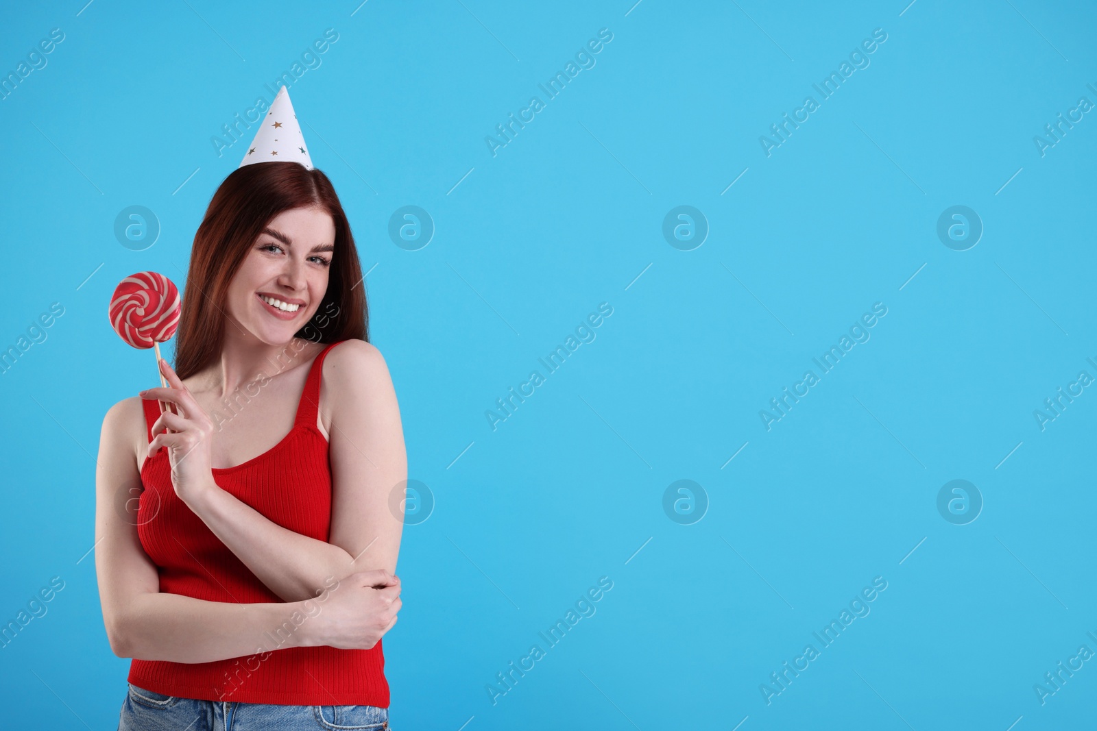 Photo of Happy woman in party hat with lollipop on light blue background, space for text