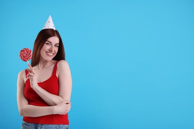 Happy woman in party hat with lollipop on light blue background, space for text