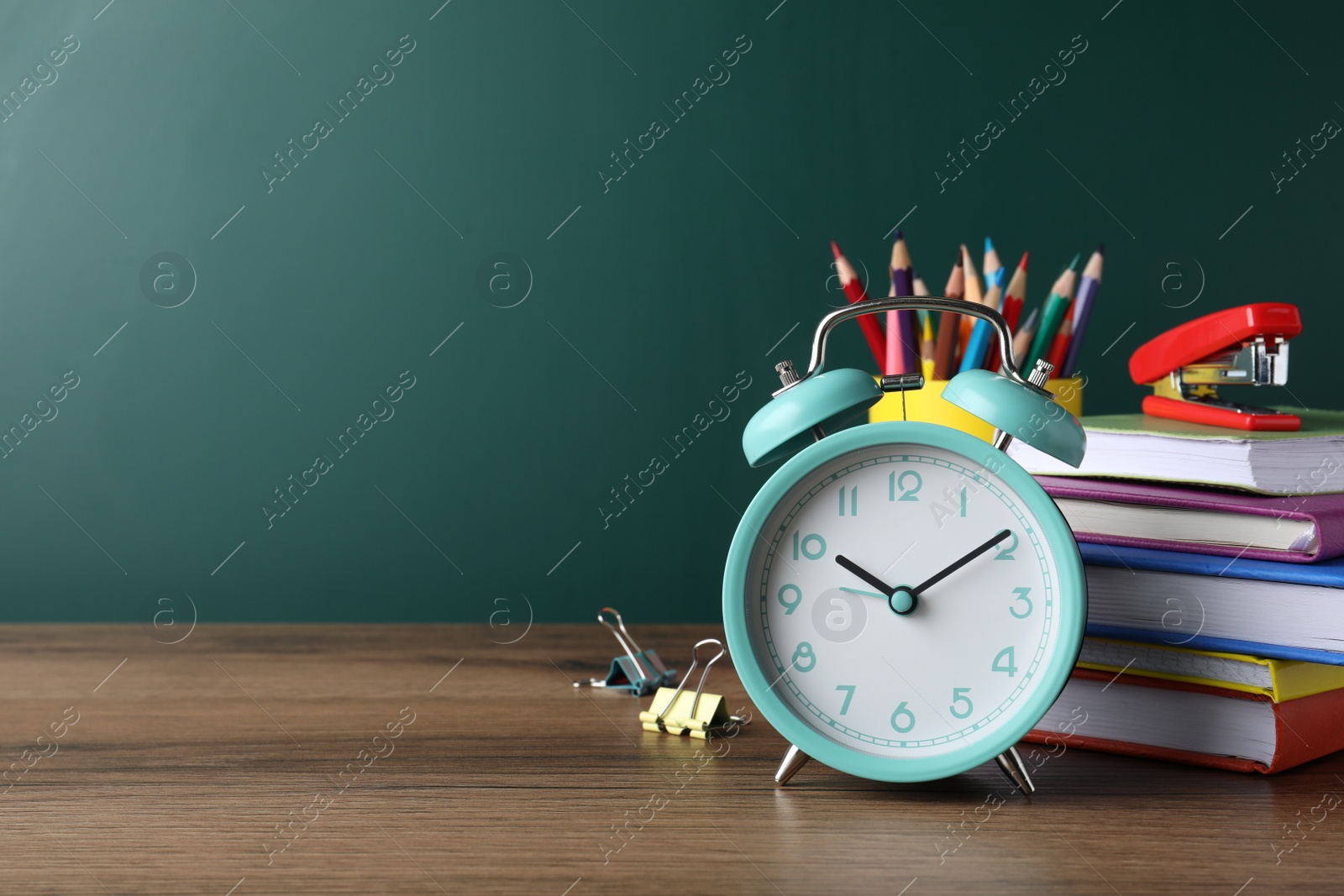 Photo of Alarm clock and different stationery on wooden table near green chalkboard, space for text. School time
