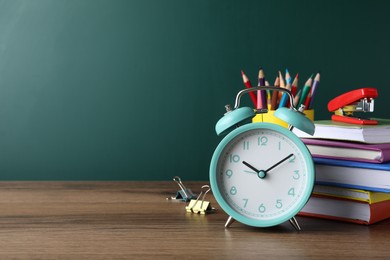 Alarm clock and different stationery on wooden table near green chalkboard, space for text. School time