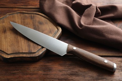 Photo of One sharp knife, cloth and board on wooden table, closeup