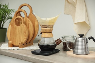 Photo of Making drip coffee. Glass chemex coffeemaker with paper filter, jar of beans and kettle on wooden countertop in kitchen