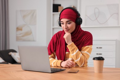 Muslim woman in hijab with headphones using laptop at wooden table in room