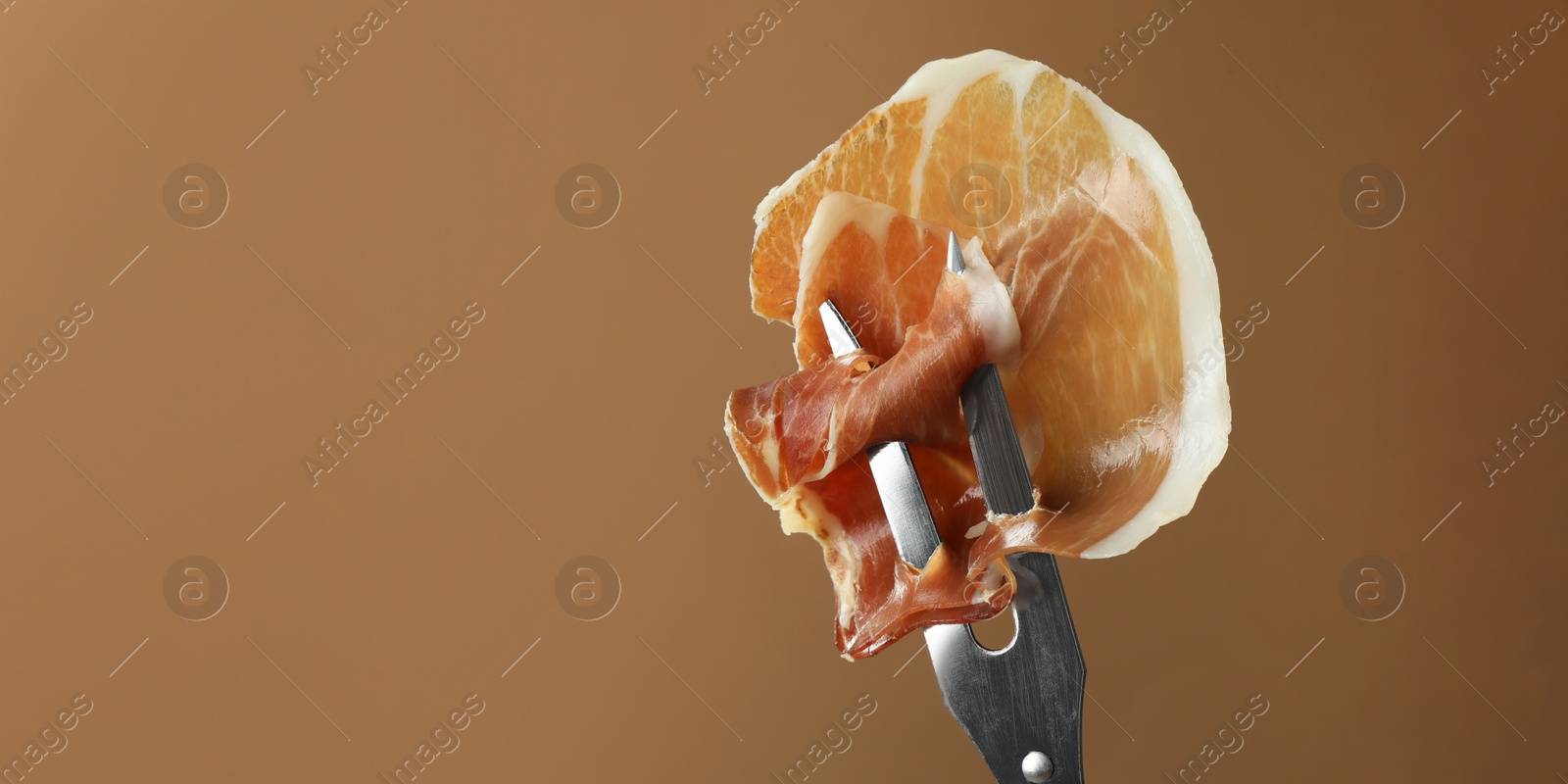 Photo of Fork with slice of tasty jamon on brown background, closeup. Space for text