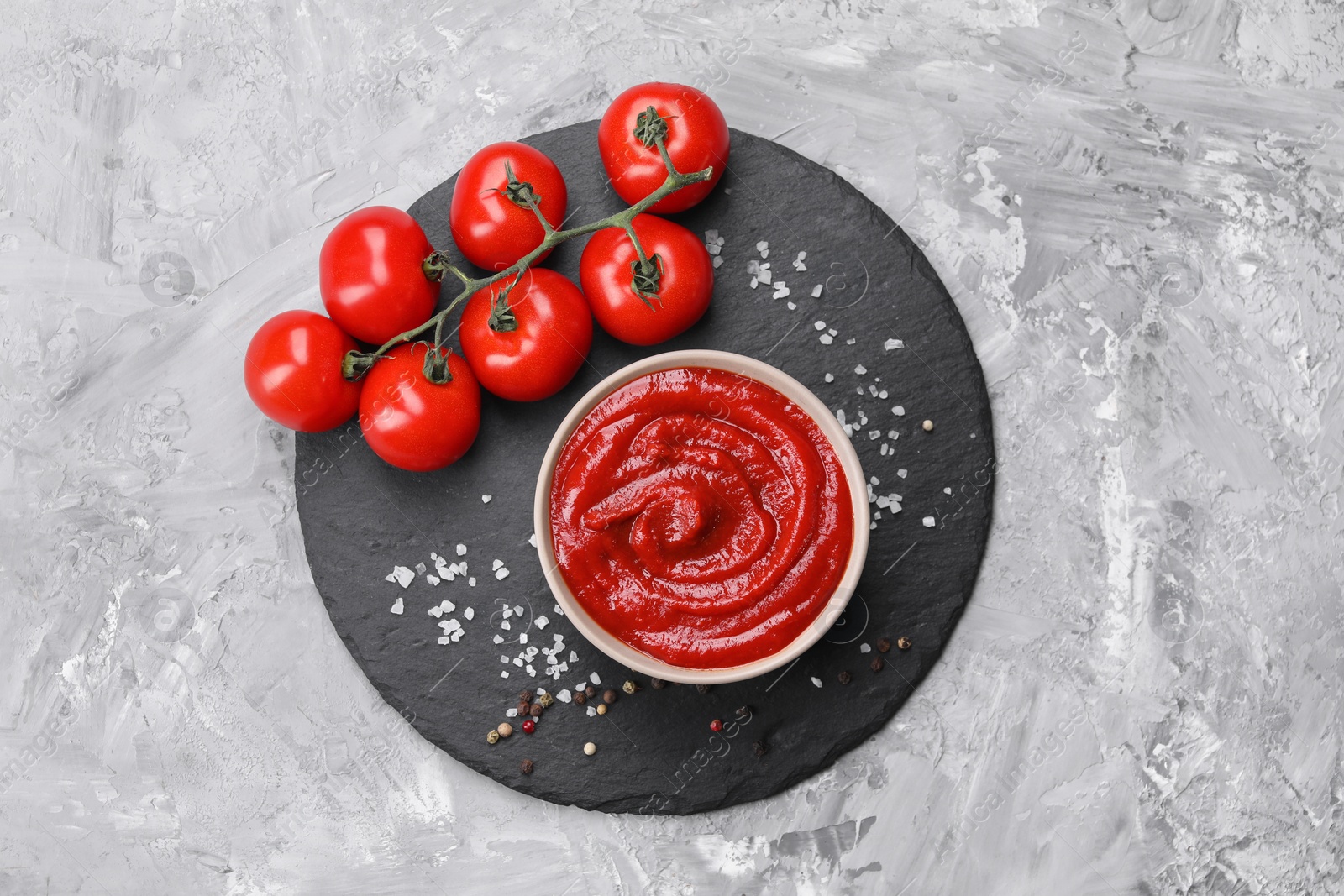 Photo of Organic ketchup in bowl, fresh tomatoes and spices on grey textured table, top view. Tomato sauce