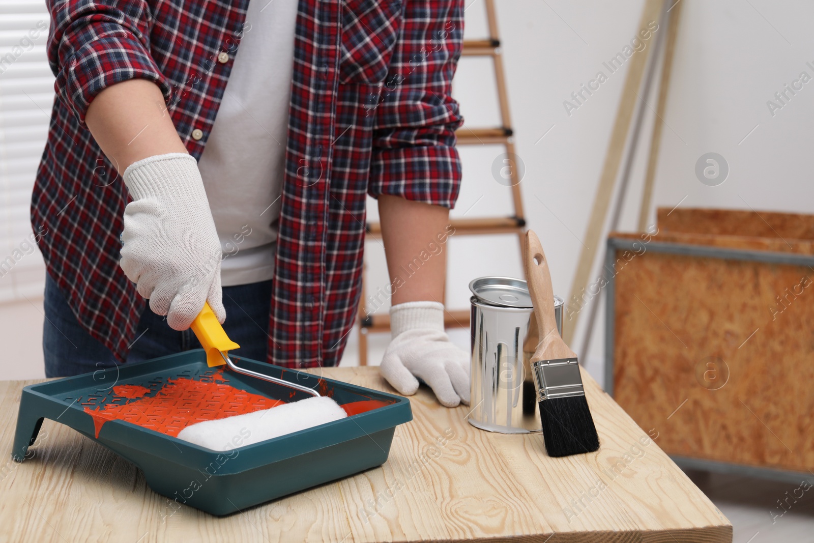 Photo of Man taking orange paint with roller from tray at wooden table indoors, closeup