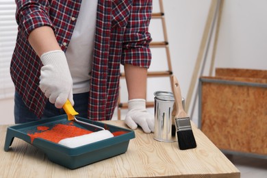 Photo of Man taking orange paint with roller from tray at wooden table indoors, closeup