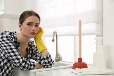 Photo of Unhappy young woman with plunger near clogged sink in kitchen