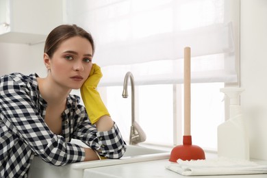 Unhappy young woman with plunger near clogged sink in kitchen