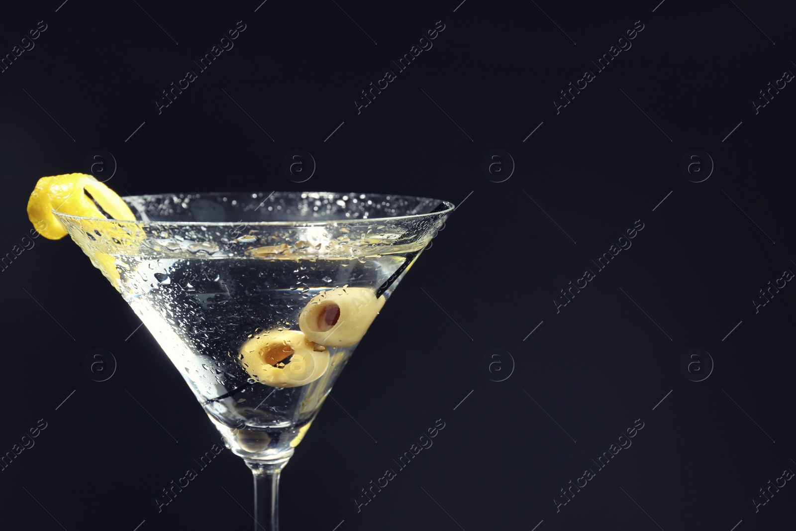 Photo of Martini cocktail with olives and lemon twist on dark background, closeup. Space for text