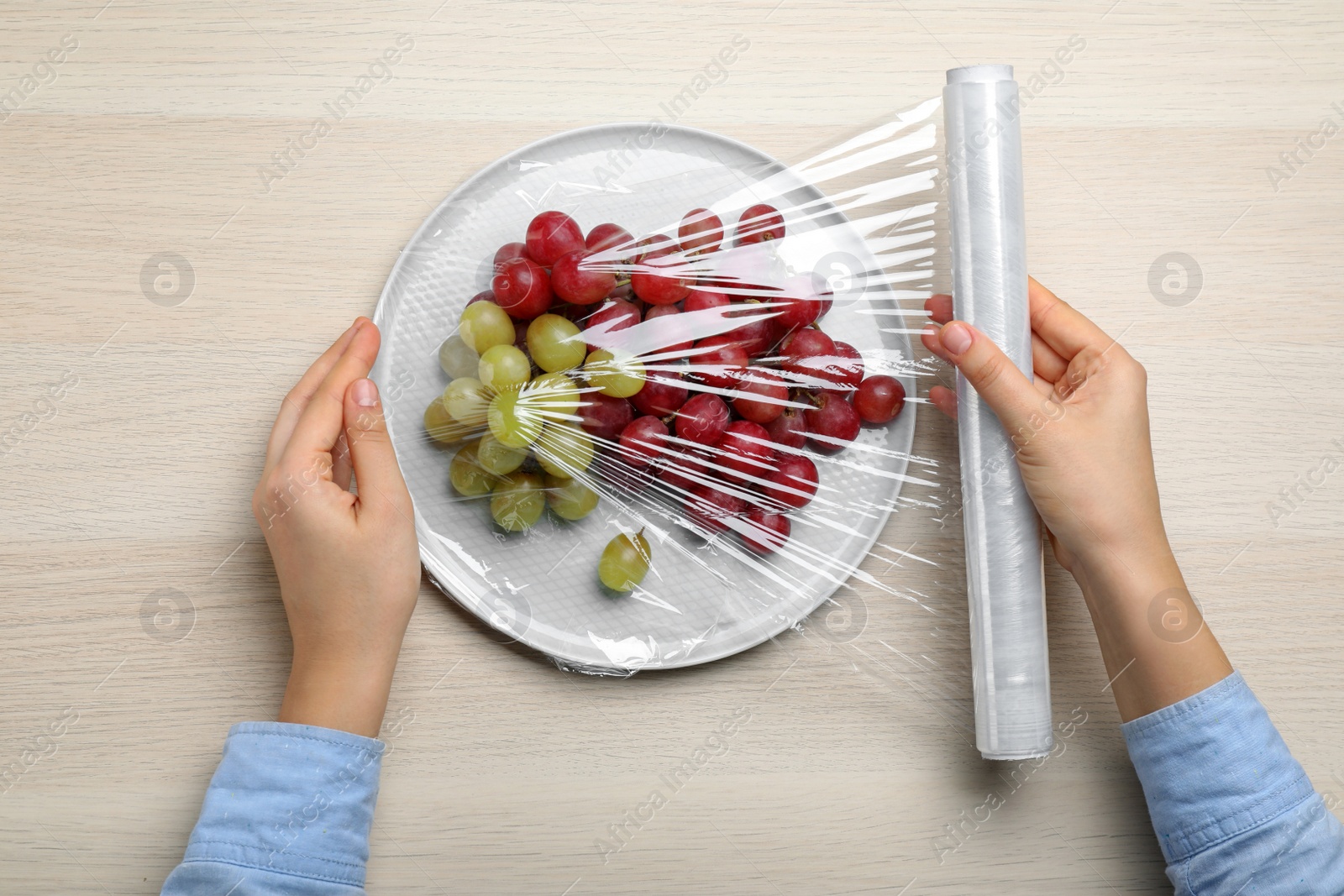 Photo of Woman putting plastic food wrap over plate of fresh grapes at wooden table, top view