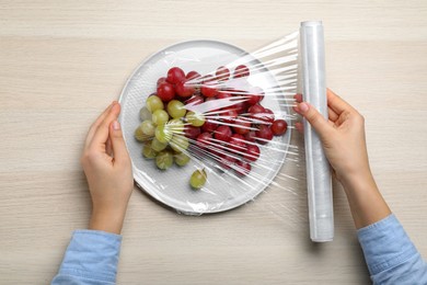 Photo of Woman putting plastic food wrap over plate of fresh grapes at wooden table, top view