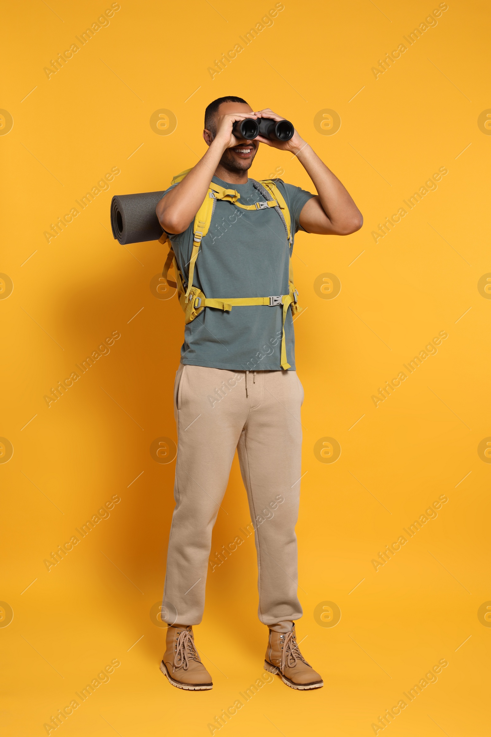 Photo of Happy tourist with backpack looking through binoculars on yellow background