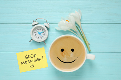 Delicious coffee, alarm clock, flowers and card with GOOD MORNING wish on light blue wooden table, flat lay