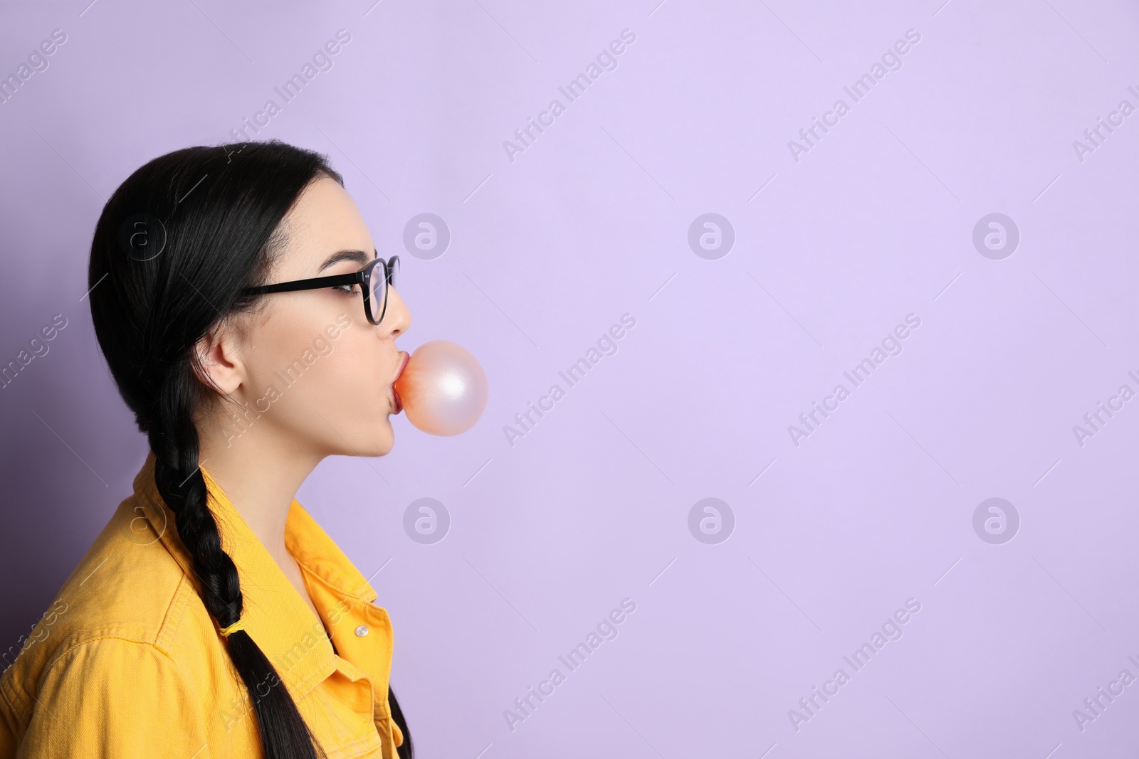 Photo of Fashionable young woman with braids blowing bubblegum on lilac background, space for text