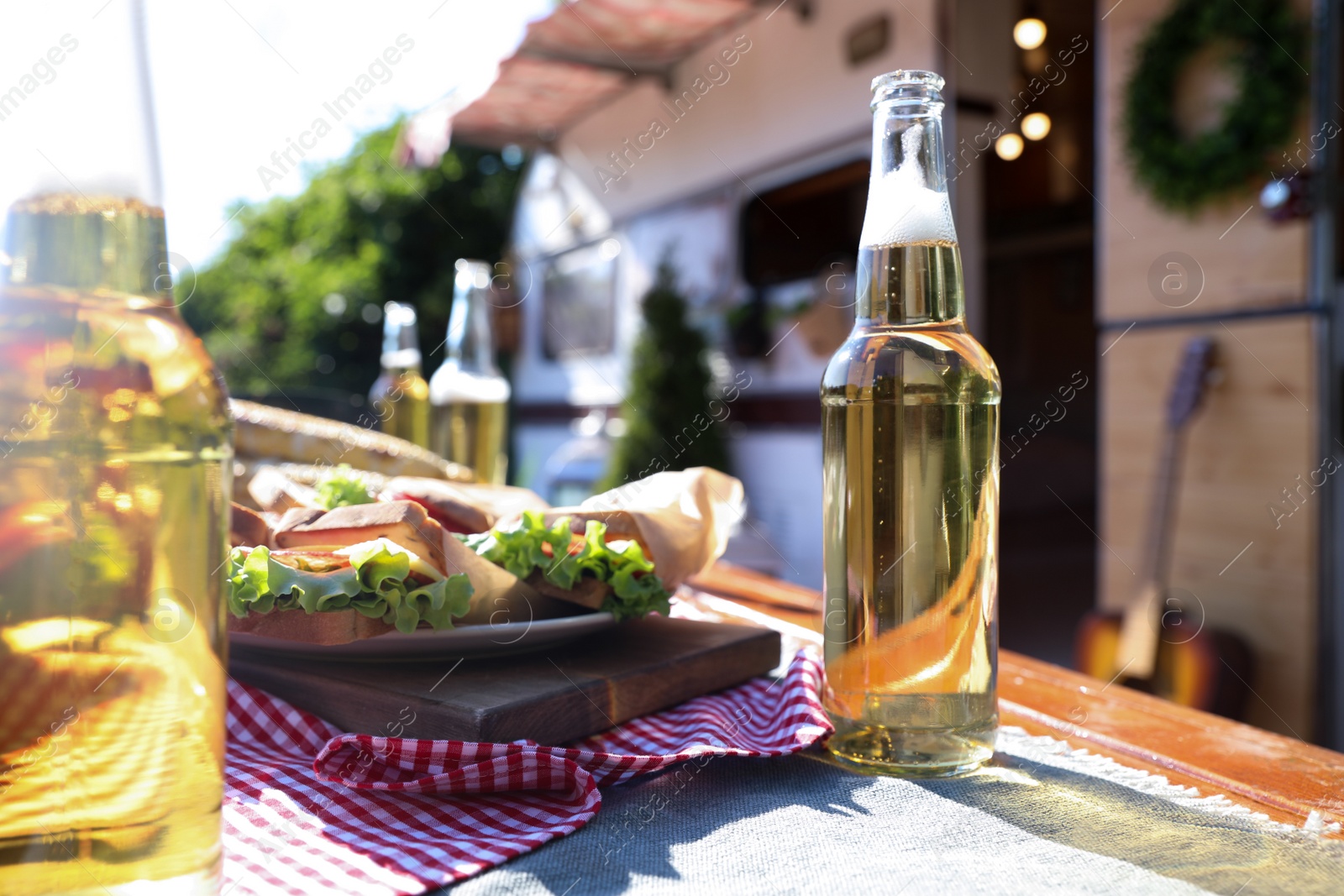 Photo of Delicious sandwiches and bottles of beer on wooden table near motorhome on sunny day. Camping season