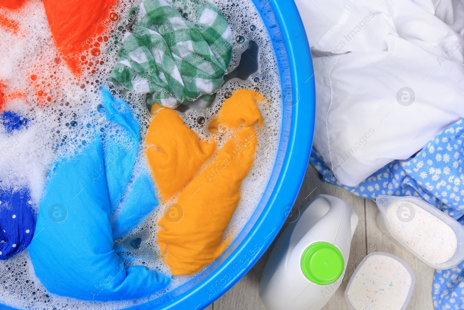 Photo of Basin with colorful clothes near bottle of detergent and powder on floor, flat lay Hand washing laundry