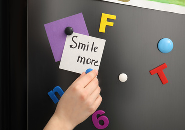 Photo of Woman putting note with phrase SMILE MORE on refrigerator door, closeup