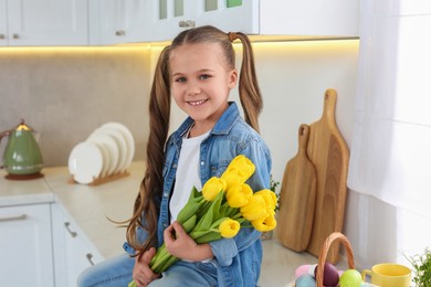 Photo of Cute girl with yellow tulips and wicker basket full of Easter eggs in kitchen