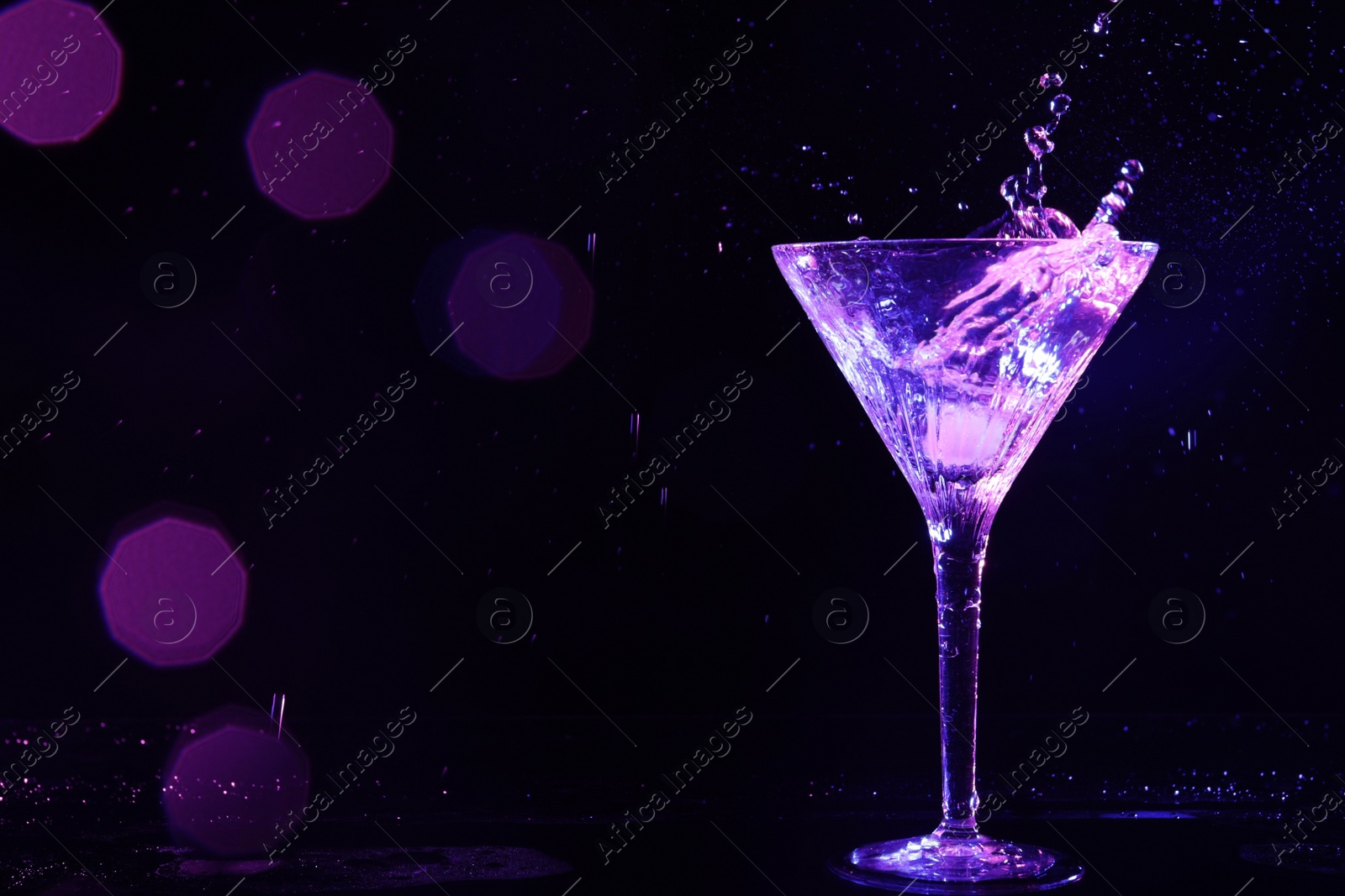 Photo of Glass of martini with ice and splashes in neon lights on dark background, bokeh effect. Space for text