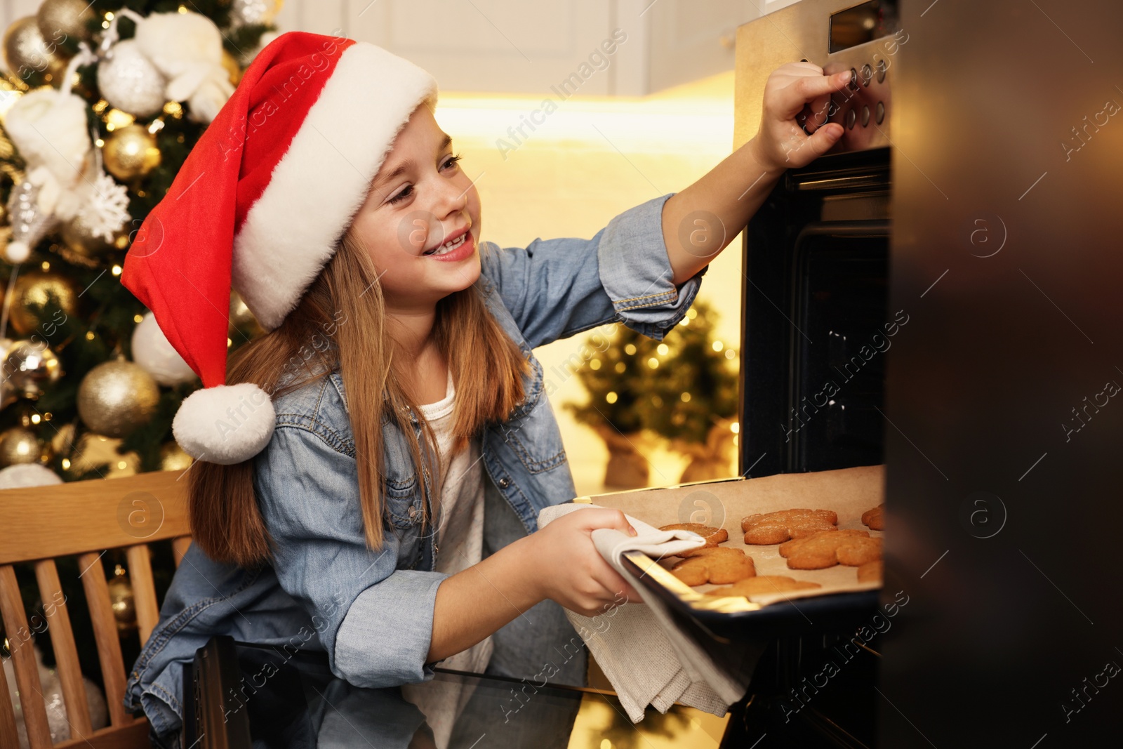 Photo of Little child in Santa hat taking baking sheet with Christmas cookies out of oven indoors