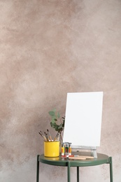 Photo of Wooden easel with blank canvas board and painting tools for children on table near color wall