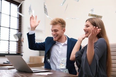 Happy young people playing online lottery using laptop under money rain in office