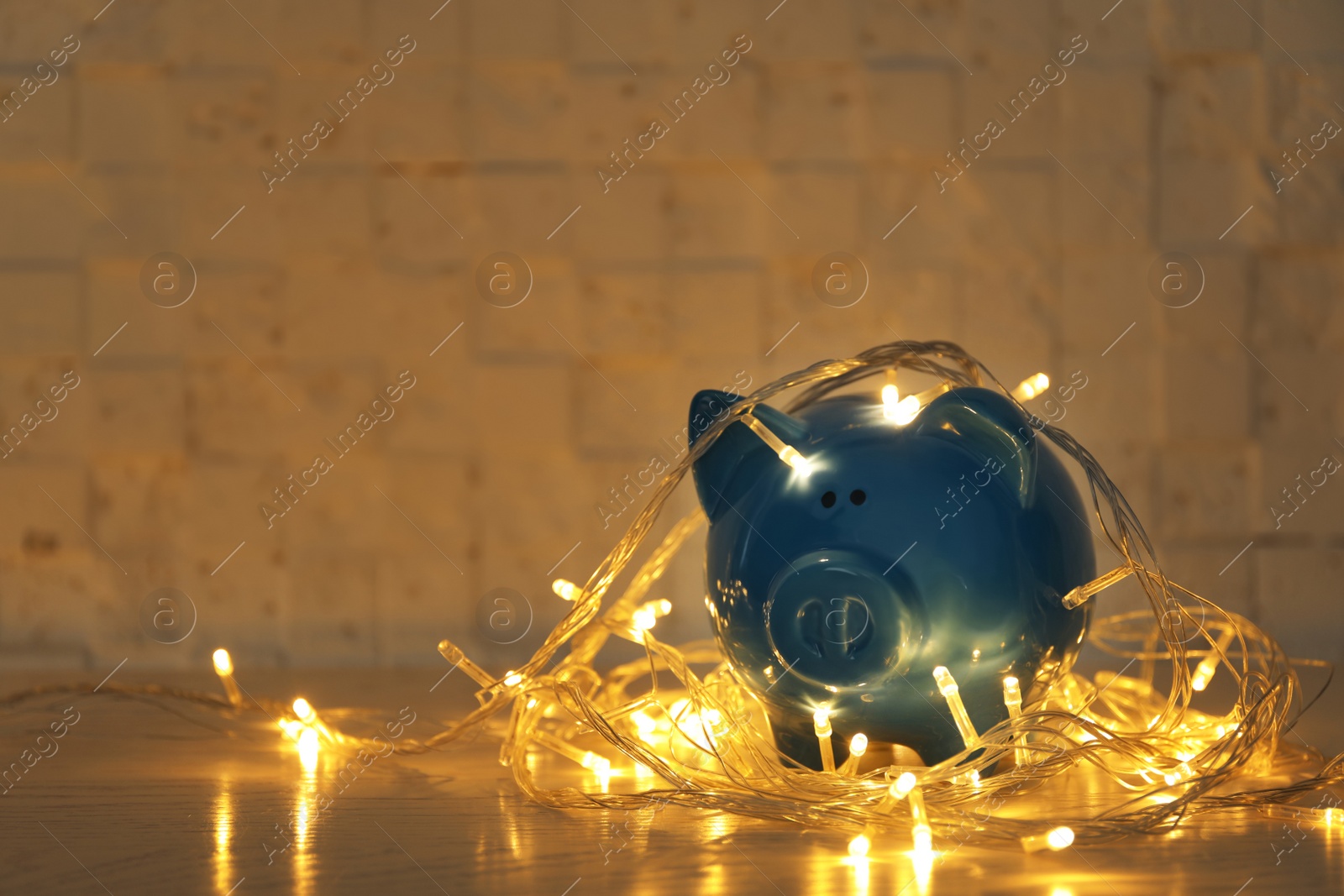 Photo of Piggy bank with Christmas lights on wooden table against textured wall. Space for text