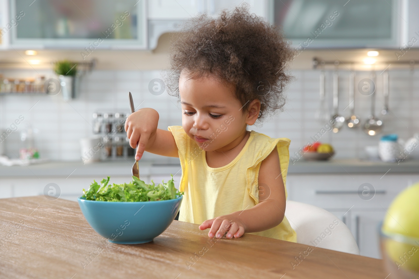 Photo of Cute African-American girl eating vegetable salad at table in kitchen