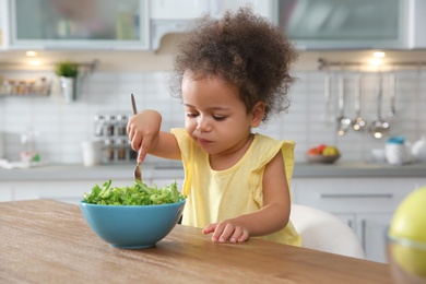 Cute African-American girl eating vegetable salad at table in kitchen