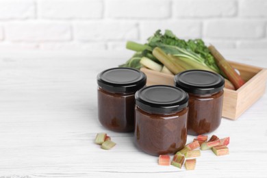 Jars of tasty rhubarb jam and cut stalks on white wooden table, space for text