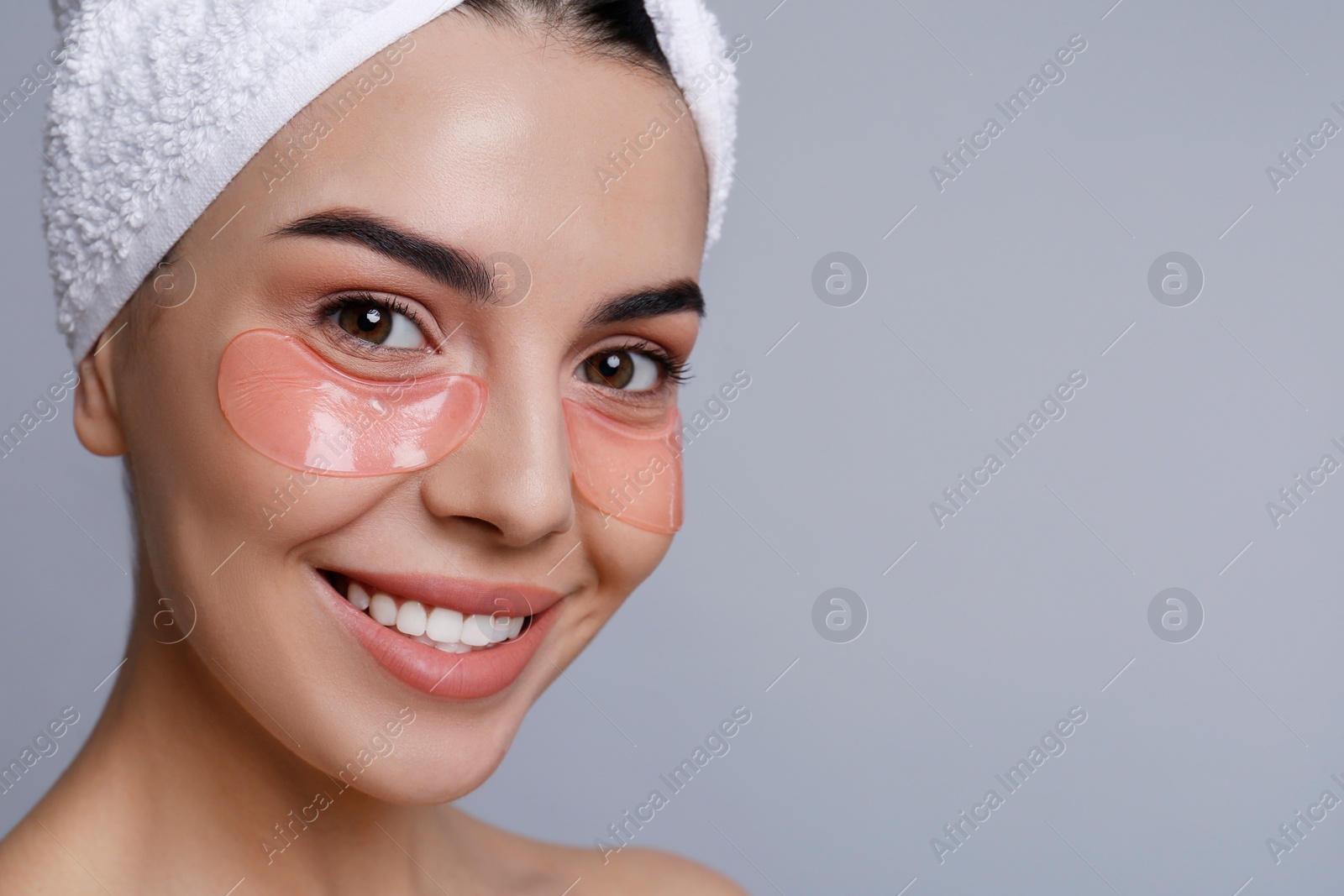 Photo of Beautiful young woman with under eye patches and hair wrapped in towel on grey background, space for text