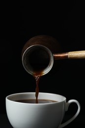 Photo of Turkish coffee. Pouring brewed beverage from cezve into cup against black background