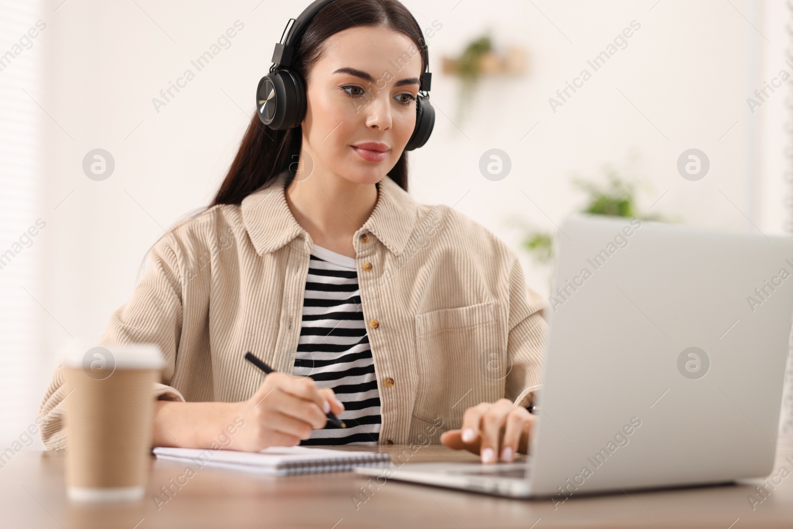 Photo of Young woman in headphones watching webinar at table in room