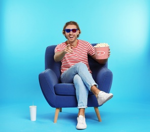 Man with 3D glasses, popcorn and beverage sitting in armchair during cinema show on color background