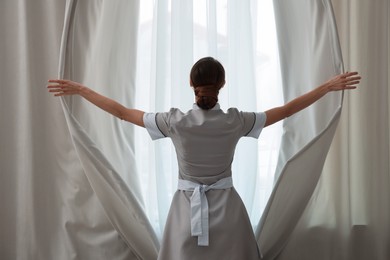 Photo of Chambermaid opening window curtains in hotel room, back view