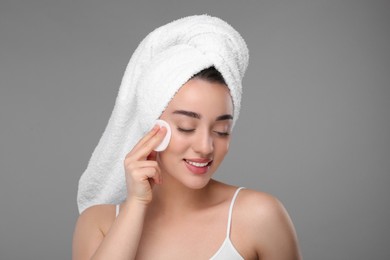 Beautiful woman in terry towel removing makeup with cotton pad on gray background