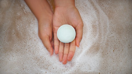 Photo of Woman holding bath bomb over water with foam, top view