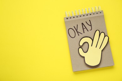 Photo of Notepad with word Okay and paper cutout of OK hand gesture on yellow background, top view. Space for text
