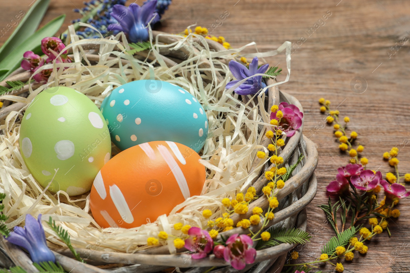 Photo of Wicker nest with painted Easter eggs and flowers on table, closeup