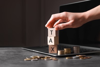 Photo of Woman with word Tax made of wooden cubes, laptop and coins at grey table, closeup. Space for text