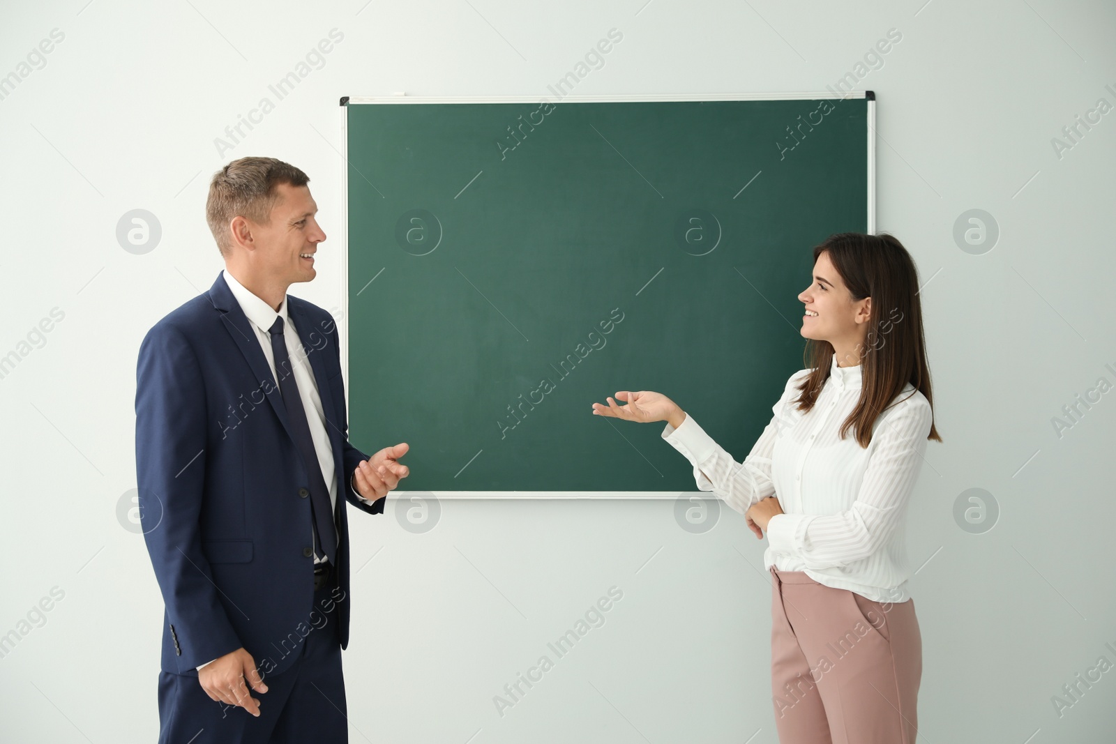 Photo of Man and woman talking near green chalkboard in classroom
